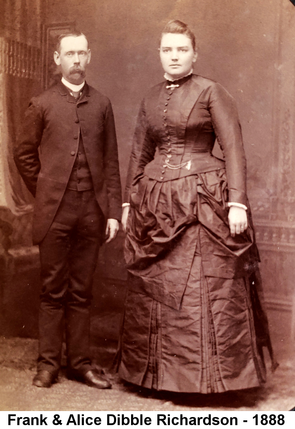 IMAGE/PHOTO: Frank & Alice Dibble Richardson - 1888: Rose-tinted full-length studio portrait photo a thin man with a bushy moustache standing next to a rather broad young woman in a dark, tightly-corseted long dress with a watch chain, the backs of their hands almost touching.